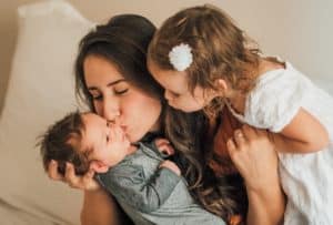 Mother kissing baby #2 with big sister watching