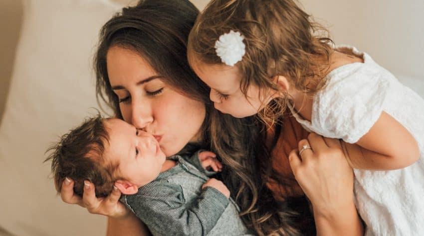 Mother kissing baby #2 with big sister watching