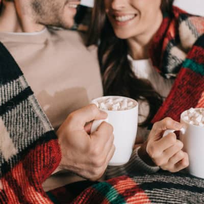 married couple snuggling with holiday blanket and sipping hot cocoa