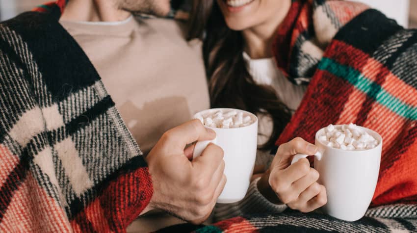 married couple snuggling with holiday blanket and sipping hot cocoa